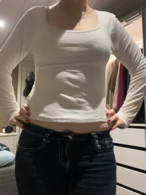 Vit långärmad topp med spetskant - Säljer en vit långärmad topp med fyrkantig halsringning och diskret spetskant runt halsen. Modellen är figurnära och har en enkel, stilren design som passar till jeans eller kjol. Materialet känns mjukt och stretchigt, perfekt för dig som gillar bekväma och snygga basplagg.