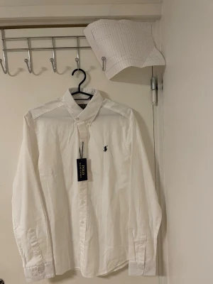 Vit skjorta  Polo Ralph Lauren - Vit skjorta från Polo Ralph Lauren med klassisk krage och broderad logga på bröstet. Skjortan har långa ärmar, knappar framtill och är gjord i mjuk bomull. Perfekt för dig som gillar stilrena och tidlösa plagg.Liknande modell som orginala. 