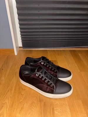 Vin röda sneakers från Lanvin - Snygga svarta sneakers från Lanvin med tåparti i matt svart skinn. Skorna har vita platta sulor och svarta snören med metallöglor. Insidan är fodrad i svart och märket syns på innersulan. Perfekt för dig som gillar stilrena detaljer. Stora i storleken