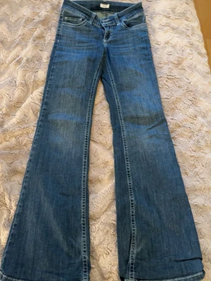 Blå bootcut jeans från Nelly - Säljer ett par blå bootcut jeans från Nelly med klassisk femficksdesign och dubbla silverfärgade knappar i midjan. Jeansen har dragkedja. Jeansen är lite söndriga vid benen 