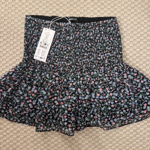Blommig kortkjol från Gina Tricot - Aldrig använt. Supersöt kortkjol från Gina Tricot med svart botten och små blommor i rosa, blått och grönt. Kjolen har smock i midjan och volangig nederdel som ger en flowy känsla. Perfekt för dig som gillar en feminin och trendig stil.