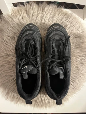 Svarta Nike Air Max 97 sneakers - Nike Air Max 97 sneakers i helsvart med vita swoosh-loggor på sidan och tungan. Skorna har en vågig design med synlig Air-sula och snörning framtill. Materialet är en mix av syntet och textil med perforerade detaljer för extra stil. Perfekt för dig som gillar streetwear och sportig vibe.