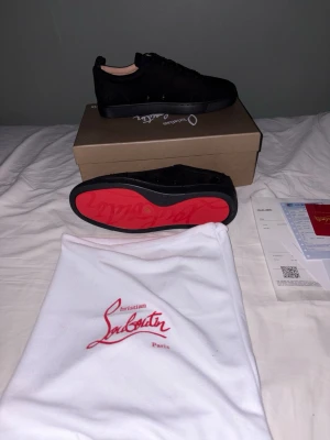 Christian Louboutin svarta sneakers - Säljer ett par svarta sneakers från Christian Louboutin i mocka med den ikoniska röda sulan. Skorna har snörning, rund tå och diskret broderad logga på hälen. Insidan är i beige läder och sulan är i gummi. Kommer med originalpåse och låda.