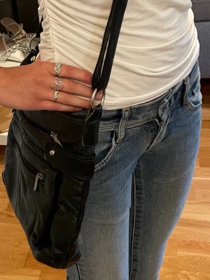 Svart axelväska i skinn med dragkedja - En svart axelväska i skinn med justerbar rem och flera praktiska fack med dragkedja.