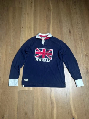 Morris marinblå rugbytröja med tryck - Säljer en marinblå rugbytröja från Morris med Union Jack-tryck och broderad logga på bröstet. Skjortan har vit krage med röd insida, vita manschetter och quiltade armbågslappar. Tillverkad i mjuk bomull och har klassisk henley-knäppning vid halsen.