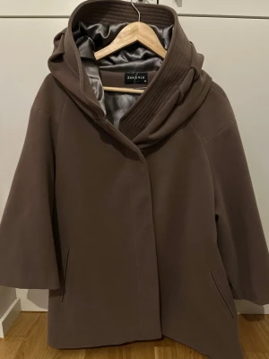Brun kappa med stor krage från Zanavle - Säljer en brun kappa från Zanavle i storlek 40. Den har en stor, snygg krage som kan stylas som huva och två fickor framtill. Insidan är fodrad med glansigt tyg i grått. Passformen är loose. Perfekt för dig som gillar stilrena och coola kappor.