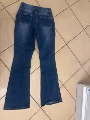 Blå bootcut jeans med slitningar - Snygga blå jeans med bootcut-snitt och slitna detaljer framtill. Jeansen har klassisk femficksdesign, knapp och dragkedja i midjan samt lätt tvättade partier för en cool look. Materialet är jeans och färgen är mörkblå med ljusare partier.