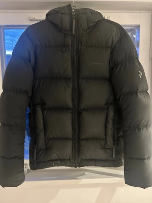 Svart dunjacka från Peak Performance - Säljer en svart dunjacka från Peak Performance med huva och dragkedja framtill. Jackan är tjock och quiltad, perfekt för kalla dagar. Klassisk design med diskret logga på bröstet och ärmen. 45cm från axel till axel (söm) Läs mer om jackan här: https://www.peakperformance.com/se/sv/product/m-rivel-jacket-g77962.html?color=52788