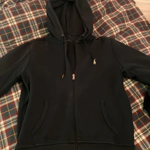 Mörkblå dress från Polo Ralph Lauren - Mörkblå dress med huva från Polo Ralph Lauren, har dragkedja framtill och två fickor. Klassisk Polo-logga broderad på bröstet. Ett litet hål vid vänster axel och lite borttvättad etikett på tjöjan men annars bra skick