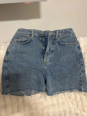 Högmidjade jeansshorts - Säljer ett par klassiska blå jeansshorts från never denim. De är högmidjade med råa, fransiga benslut och har knappar framtill. Shortsen har fem fickor och bälteshällor. 