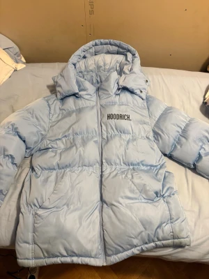 Ljusblå pufferjacka från Hoodrich - Ljusblå pufferjacka från Hoodrich med stor logga på både bröstet och ryggen. Jackan har huva, dragkedja framtill och två stora fickor. Insidan har coola axelremmar med H-mönster för att bära jackan som en ryggsäck. Perfekt för kalla dagar.