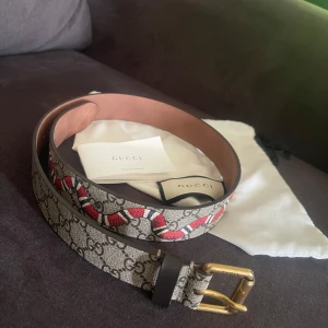 Gucci bälte med orm och GG-mönster - Säljer ett Gucci bälte med klassiskt GG-mönster i beige och brunt, dekorerat med en röd och vit orm längs bandet. Bältet har guldfärgat spänne och detaljer i svart läder. Perfekt accessoar för att lyfta vilken outfit som helst.