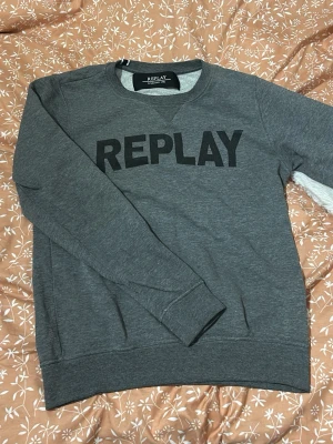 Grå sweatshirt från Replay - Säljer en grå sweatshirt från Replay med stor svart logotyp på bröstet. Tröjan har rund hals, långa ärmar och ribbade muddar vid ärmslut och nederkant. Perfekt för chill dagar och enkel att matcha med jeans eller joggers.