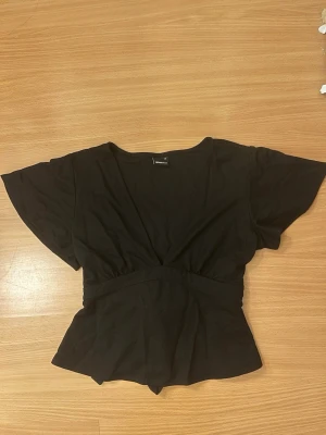 Svart omlottopp från Gina Tricot XS - Säljer en svart omlottopp från Gina Tricot i storlek XS. Toppen har korta ärmar, v-ringad hals och ett knytband i midjan som ger en snygg siluett. Materialet är mjuk bomull och modellen är croppad med lätt volang nedtill.