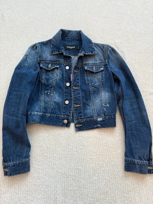 Blå jeansjacka från DSQUARED2 - Snygg blå jeansjacka från DSQUARED2 med coola slitningar och färgstänk. Klassisk krage, långa ärmar och silverfärgade knappar framtill. Jackan har två bröstfickor med lock och är croppad i modellen. Perfekt för dig som gillar streetstyle. Liten i storleken. 