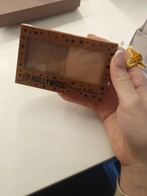 W7 Hollywood Bronze & Glow duo - Säljer en W7 Hollywood Bronze & Glow duo med bronzer och highlighter i en brun plastförpackning med stjärnmönster. Bronzer är varmbrun och highlightern är ljus champagnefärgad. Paletten har en inbyggd spegel och är perfekt för att skapa glow och definition.