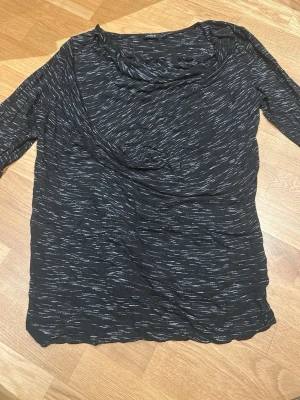 Svart mönstrad topp från Lindex - Säljer en svart långärmad topp från Lindex i storlek M. Toppen har ett diskret randigt mönster i vitt och en lösare passform med draperad ringning. Materialet är mjukt och stretchigt, perfekt för en chill och snygg look.