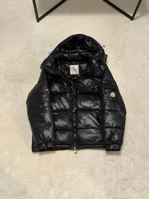 Svart pufferjacka från Moncler - Svart pufferjacka från Moncler med glansig finish, huva och dragkedja framtill. Jackan har två sidofickor med dragkedja och en extra ficka på ärmen med Moncler-logga. Perfekt för kalla dagar och har en riktigt snygg streetstil.