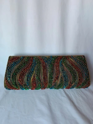 Färgglad aftonväska från Oscar & Clothilde - Säljer en unik kuvertväska från Oscar & Clothilde med broderade pärlor i blått, grönt, rött och guld. Väskan har ett vågigt mönster och stängs med magnetlås. Insidan är svart med en liten ficka. Aldrig använd så i absolut toppskick. Extra pärlor medföljer. 23-26 cm lång, 10 cm hög. 