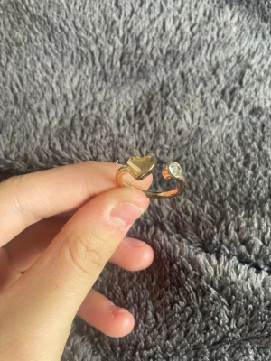 Guldfärgad ring med hjärta och sten - Snygg guldfärgad ring med öppet band, dekorerad med ett litet hjärta i metall på ena sidan och en klar sten på andra sidan. Ringen har en modern och enkel design som passar perfekt för att ge din stil lite extra glans. Den är justerbar och super fin!🤍