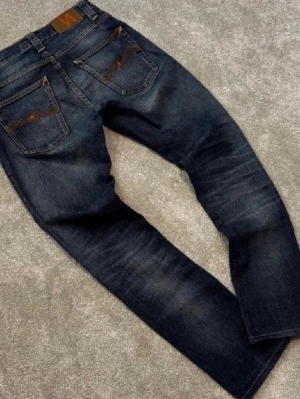 Nudie Jeans || Grim Tim - Snygga mörkblå Grim Tim jeans från Nudie Jeans med klassisk femficksdesign och orangea sömmar. Jeansen är tillverkade i slitstarkt denim med coola slitningar på framsidan och bakfickor med Nudie-signatur. Perfekt för dig som gillar en tidlös look. W28 L32