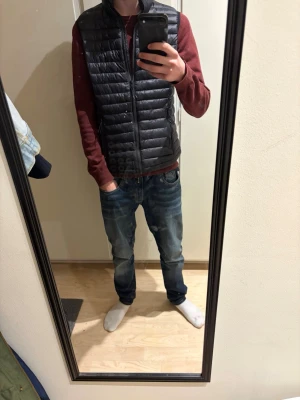 Svart dunväst med dragkedja - Säljer en svart dunväst med horisontella quiltade sömmar och hög krage. Västen har två fickor med dragkedja och stängs med en dragkedja framtill. Perfekt att slänga över en hoodie eller tröja för extra värme.