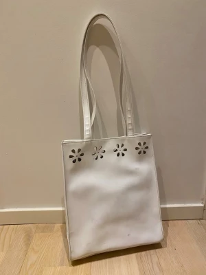 Vit shoppingväska med blommiga hål - Säljer en vit väska med långa axelband och blommiga utskärningar upptill. Insidan har rutigt foder och en  innerficka med dragkedja. Stängs med magnetknapp.