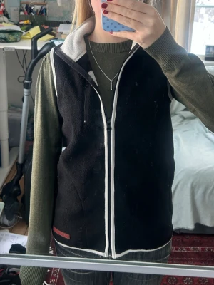Svart fleeceväst med vit kant - Svart fleeceväst med vit dragkedja och vita detaljer längs kanterna. Hög krage och praktiska fickor med dragkedja. Perfekt att slänga på över en hoodie eller tröja för extra värme. Enkel och stilren design.