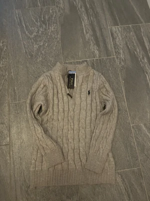 Grå stickad tröja Polo Ralph Lauren - Snygg grå stickad tröja från Polo Ralph Lauren i storlek S. Tröjan har kabelstickat mönster, ribbade muddar och en liten broderad logga på bröstet. Perfekt för dig som gillar klassisk och stilren design.