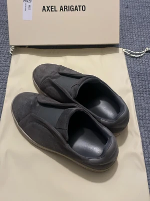 Grå mocka slip-on sneakers Axel Arigato - Säljer ett par grå slip-on sneakers från Axel Arigato i mocka med elastisk detalj över vristen. Skorna har rund tå, vit platt sula och diskret logga på sidan av sulan samt på plösen. Perfekta för dig som gillar minimalistisk och clean stil.