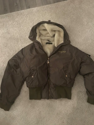 Brun pufferjacka från Baby Phat - Säljer en mörkgrön pufferjacka från Baby Phat med guldig brodyr och logga på bröstet och ryggen. Jackan har mysigt vitt foder, huva och coola detaljer som dragkedja med logga och fickor med knappar. Ribbad mudd nedtill och vid ärmslut för extra komfort. köptes för 945.