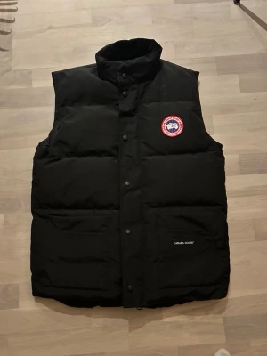 Svart dunväst från Canada Goose - Säljer en svart dunväst från Canada Goose med hög krage och tryckknappar framtill. Västen har två stora fickor och den klassiska Canada Goose-loggan på bröstet. Perfekt för kalla dagar när du vill hålla stilen. Passar M och L
