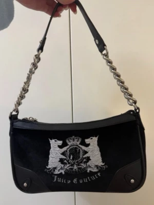 Juicy Couture svart axelväska - Juicy Couture axelväska i svart sammet med silverfärgade detaljer och kedjerem. Väskan har broderat emblem framtill och dragkedja upptill. Insidan är fodrad med logotypmönster och har en innerficka. Medföljer en liten silverfärgad necessär, svart rosett och hjärtformat tag.