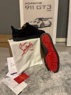 Mörkblå Christian Louboutin sneakers - Säljer ett par mörkblå sneakers från Christian Louboutin i mocka med den ikoniska röda sulan och diskret logga på sidan. Skorna har snörning och en stilren, rund tå. Kommer med originalpåse och box. Perfekta för dig som vill ha något exklusivt och tidlöst.