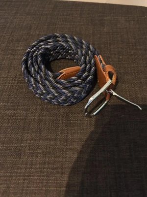 Blå flätad bälte med läderdetaljer - Säljer ett snyggt blått flätat bälte med bruna läderdetaljer och silverfärgat spänne. Bältet har en cool mix av mörkblå och svarta toner i flätningen och passar perfekt till jeans eller chinos för en avslappnad men stilren look.