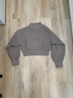 Grå croppad stickad tröja - Säljer en grå stickad tröja med ballongärmar och ribbade muddar. Tröjan är cropped men når relativt långt ner över midjan ändå. Tröjan har en liten reva på sidan av midjan men som hållit sig stabil under en längre tid.