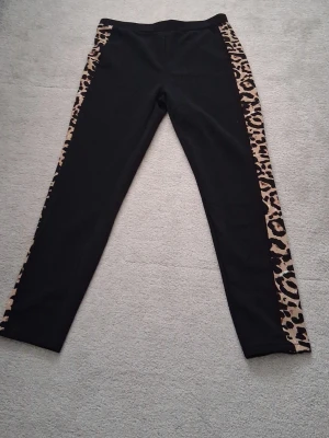 Svarta leggings med leopardmönster SHEIN - Säljer ett par svarta leggings från SHEIN i storlek XL med breda leopardmönstrade ränder längs båda sidorna. Stretchigt material som sitter tight och ger en cool kontrast mellan svart och beige/brunt. Perfekta för dig som vill sticka ut lite extra.