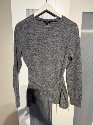 Grå långärmad topp med knyt Happy Holly - Mysig gråmelerad långärmad topp från Happy Holly i storlek 36/38. Modellen har ett snyggt knyt i midjan som ger en fin siluett. Perfekt till både jeans och kjol för en chill men ändå stilsäker look.