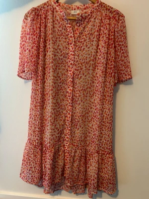 Mönstrad kortklänning från JDY - Söt kortklänning från JDY med röd- och rosa mönster på beige botten. Klänningen har volangkant nertill, knappar framtill, korta ärmar och ett knytband i midjan. Luftigt material som känns perfekt för varma dagar.