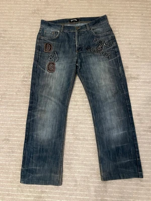 Dolce & Gabbana blå jeans med brodyr - Snygga blå jeans från Dolce & Gabbana med stora broderade D&G-loggor och text på både fram- och baksidan. Jeansen har klassisk femficksdesign, raka ben och coola slitningar. Materialet är jeans i bomull och detaljerna sticker verkligen ut.
