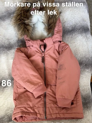 Rosa parkajacka med fuskpäls - Snygg rosa parkajacka med huva och fuskpäls. Jackan har dragkedja framtill och ribbade muddar vid ärmslut. Perfekt för kyliga dagar och har en cool, lite längre passform. Färgen är en mjuk rosa ton och huvan ger extra stilpoäng.🌸 Mörkare/fläckar på vissa ställen efter lek. Storlek 86