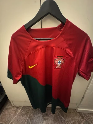 Portugal Ronaldo 7 Nike fotbollströja - Säljer en Portugal landslagströja från Nike med Ronaldo 7 på ryggen. Tröjan är röd med gröna detaljer och gula tryck, har FIFA World Cup Qatar 2022 och Living Football FIFA patchar på ärmarna. Tillverkad i polyester med Dri-FIT teknologi för extra komfort.