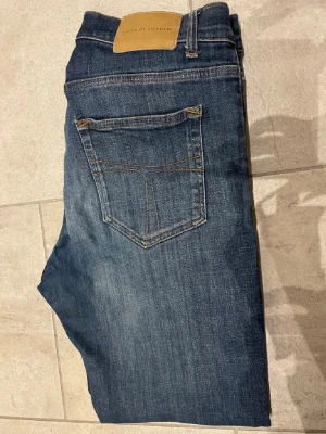 Tiger of Sweden blå jeans 32/34 - Snygga blå jeans från Tiger of Sweden i klassisk rak modell. Jeansen har fem fickor, guldfärgade sömmar och en läderpatch bak i midjan. Tillverkade i Italien av slitstarkt denimtyg. Perfekta för dig som gillar stilrena och tidlösa jeans. Storlek 32/34
