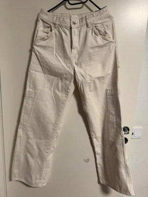 Beiga cargopants med raka ben - Säljer ett par beiga cargopants med raka ben och stora fickor på sidorna. Byxorna har normal midja, bälteshällor och är gjorda i ett slitstarkt bomullsmaterial. Perfekta för en chill och avslappnad stil. Små i storlek 