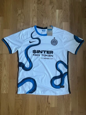 Inter Milan Nike fotbollströja vit/blå - Inter Milan bortatröja från Nike i vitt med blå och svart orm-mönster. Tröjan har blå detaljer vid krage och ärmslut, samt tryck med klubbmärke och sponsorloggor. Perfekt för dig som älskar fotboll och vill sticka ut på planen.