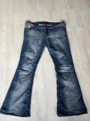 True Religion blå bootcut jeans, stl 32 - Säljer ett par blå jeans från True Religion med tydliga röda sömmar och ikoniska bakfickor. Modellen har en bred bootcut och klassisk femficksdesign. Jeansen är tillverkade i slitstarkt denim och har en snygg tvättad look.Använda några gånger och jag säljer dessa på grund ut av att dom är för stora för mig och därför ingen användning av längre.Jag skulle nog säga att dom passar någon som är mellan L-XL