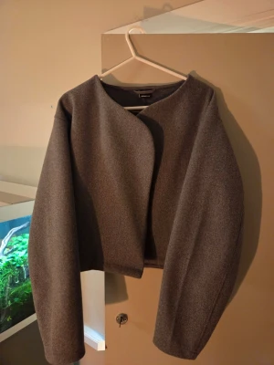 Grå omlott kappa med halsduk - Säljer en stilren grå omlott kappa från Gina Tricot med en halsduk. Jackan har en croppad passform, lång ärm och minimalistisk design utan knappar. Materialet är mjukt och känns som ullblandning. Perfekt för dig som gillar clean och modern look.