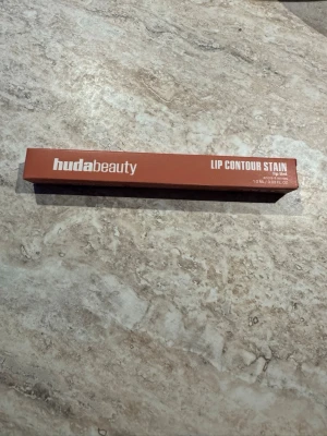 Huda Beauty Lip Contour Stain Soft Nude - Lip Contour Stain från Huda Beauty i färgen Soft Nude. Kommer i en smal, rektangulär förpackning i en varm nude nyans. Perfekt för att skapa definierade och naturliga läppkonturer. Innehåller 1 ml lip tint.