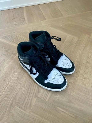 Nike Air Jordan 1 svart/vit sneakers - Säljer ett par klassiska Nike Air Jordan 1 sneakers i svart och vitt skinn. Skorna har högt skaft, svart snörning och den ikoniska Air Jordan-loggan på sidan. Perfekta för dig som vill ha en tidlös och streetinspirerad look.