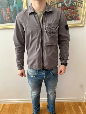 Stone Island overshirt  - Säljer en grå overshirt från Stone Island med dragkedja framtill och stor bröstficka med knapp. Klassisk patch på vänster ärm och elastiska muddar. Perfekt till lager-på-lager och riktigt clean look. Grå aktig 